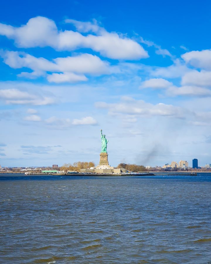 A view of the Statue of Liberty that's in the middle of the photo and is close enough to see that there's people on the island. In front of the statue is lots of water and behind you can see more plants and buildings on the land "Non-Touristy Activities for Return Visitors to New York City"