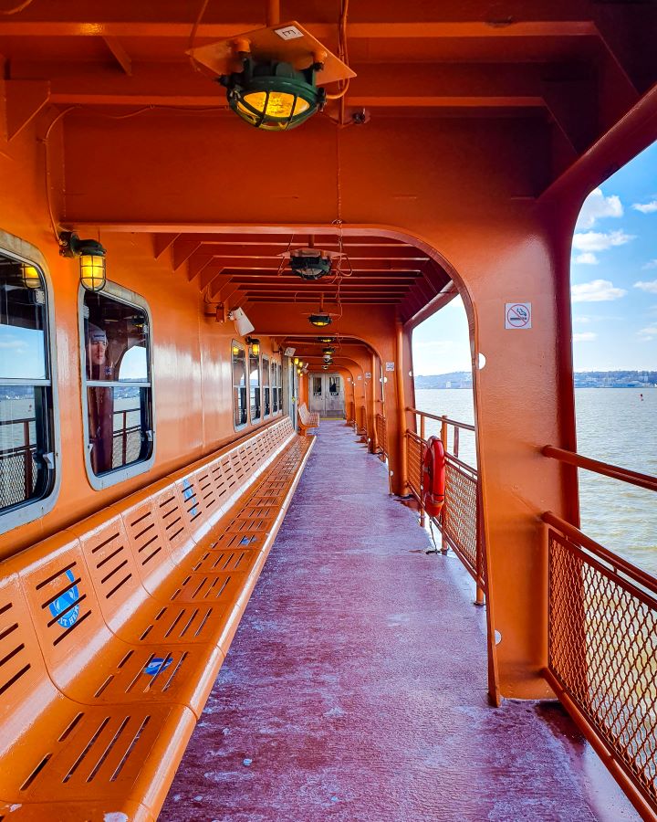 The outdoor section of the Staten Island Ferry in New York City, USA. The whole thing is a bright orange colour and you can see the water on the right. On the left is outdoor seating and there's a walkway leading to a door inside the ferry. Over the walkway and seating is the orange roof with some lights on it too and there's no other people around "Non-Touristy Activities for Return Visitors to New York City"