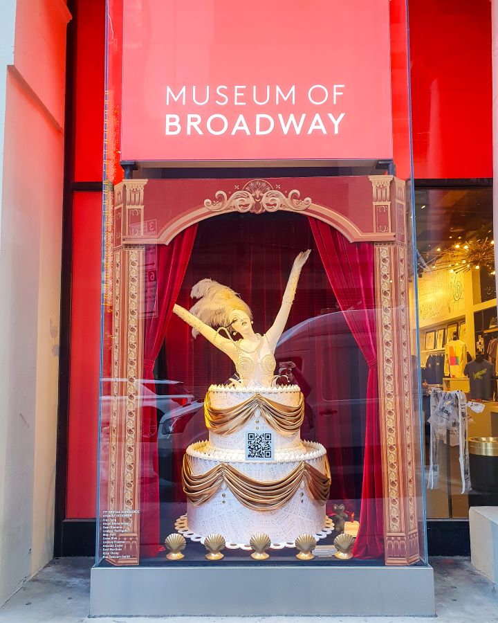 Outside the Museum of Broadway in New York City, USA. There's a red sign saying "Museum of Broadway" above a mannequin of a woman popping out of a cake with a feathered head piece "The Ultimate Guide for Musical Theatre Fans In New York"