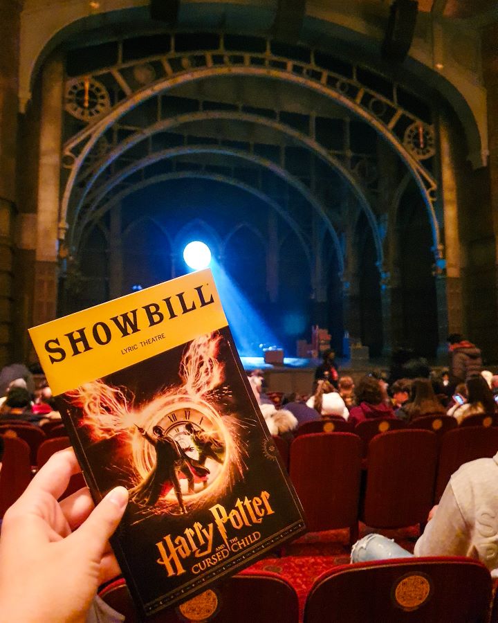 A hand holding a little book that says "Showbill" up the top in a yellow box and "Harry Potter and the Cursed Child" on the bottom in New York City, USA. Behind the Playbill are red seats and a stage with a circle spotlighting the stage and 4 big archways above the stage "The Ultimate Guide for Musical Theatre Fans In New York"