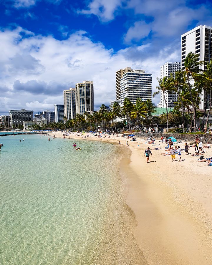 Looking at Waikīkī beach in O'ahu, Hawaii. On the right is the yellow sand, on the left is the clear blue water. There's lots of people on the beach, palm trees and behind the beach is lots of high rise buildings. "How to See O'ahu Without a Car"