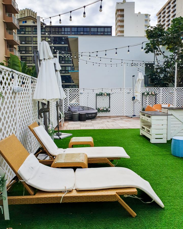 Inside the Beach Waikiki Boutique Hostel where we stayed in Waikīkī, O'ahu, Hawaii. The photo was taken in the outdoor section of the hostel so there are two deck chairs, some umbrellas, some fake grass and some more furniture. There's also fairy lights over the area and some more high rise buildings in the background. "How to See O'ahu Without a Car"