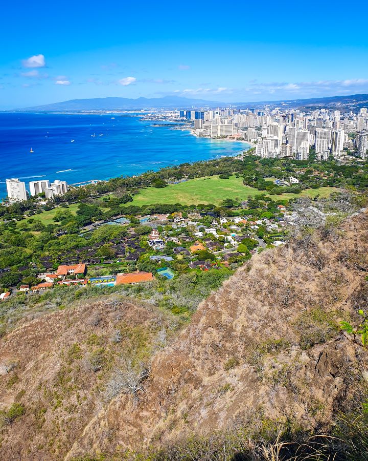 Looking down on Waikīkī in O'ahu, Hawaii. The bottom of the photo is the brown hill and some green fields and trees at the bottom of the hill with houses with orange roofs. After the fields and trees are the high rise buildings and on the left is the blue ocean with big hills in the far distance. "How to See O'ahu Without a Car"