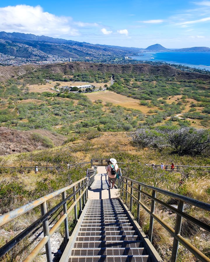 Looking down into the Diamond Head crater in O'ahu, Hawaii. There's a set of stairs leading down with people on them. Behind the stairs is a big open brown space with lots of green trees and the crater walls can be seen in a semi circle. Beyond that are more hills, and the blue water. "How to See O'ahu Without a Car"