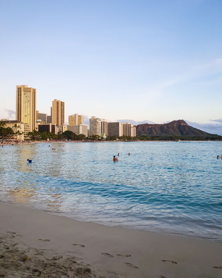 On Waikīkī beach in O'ahu, Hawaii, looking across the water at some high rise buildings and a big hill on the right which is Diamond Head. There are people in the water and footprints in the sand in the foreground and the light is an early sunset light. "How to See O'ahu Without a Car"