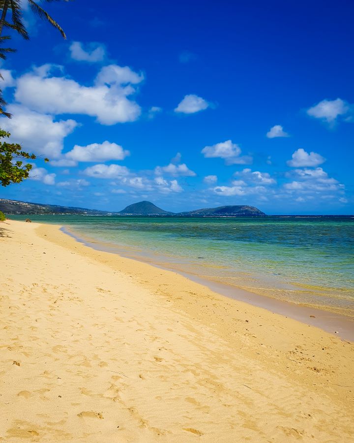 Another beautiful beach which is Kahala Beach in O'ahu, Hawaii. The left of the photo is yellow sand with the tips of some green trees, and on the right is blue and green water and in the distance are some hills including Koko Head. Above is a blue sky with some small clouds. "How to See O'ahu Without a Car"