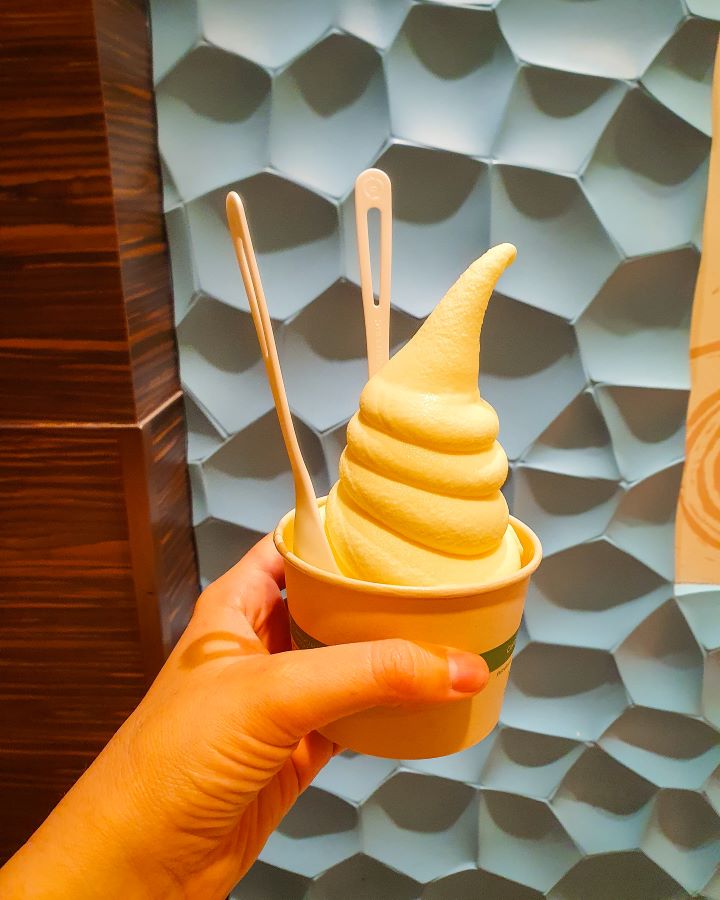 A hand holding a cup with a big spiral of yellow/white soft serve inside. There's also two spoons in it and behind it is a blue and brown wall. "How to See O'ahu Without a Car"